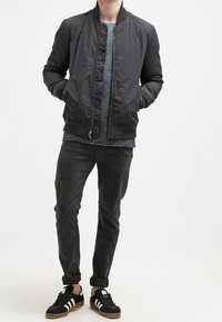Giubbotto bomber nero con zip frontale, indossato sopra una maglietta grigia con texture, abbinato a jeans neri strappati e sneakers nere con strisce bianche.
