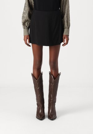 Persona que lleva botas de cowboy de cuero marrón oscuro con punteras puntiagudas y costuras decorativas, combinadas con una falda negra y mangas largas de cuadros.