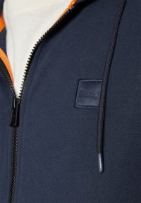 Marine blauwe hoodie met ritssluiting en oranje accenten, voorzien van een gestructureerde stof, een logo-patch op de borst en een zwarte rits met een stoffen trekker.