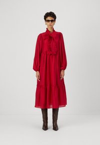 Piros, hosszú ujjú ruha fodros gallérral és réteges szoknyával. Könnyű anyagból készült, ballonujjakkal és laza fazonnal rendelkezik. Barna, térdig érő csizmák.