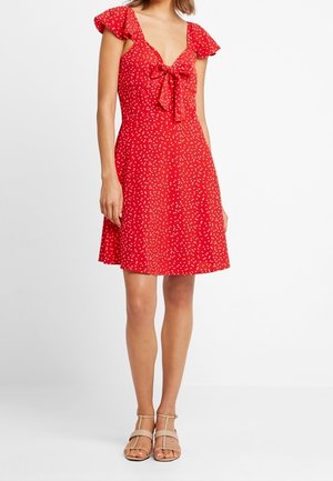 Femme portant une robe rouge à manches courtes, longueur genou, à pois blancs et un nœud sur le devant, associée à des sandales beige à bouts ouverts.