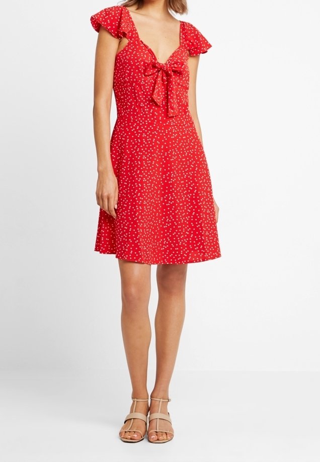 Femme portant une robe rouge à manches courtes, longueur genou, à pois blancs et un nœud sur le devant, associée à des sandales beige à bouts ouverts.