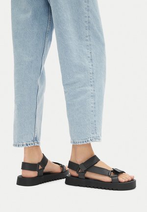 Sandalias planas negras con múltiples correas ajustables, acabado mate texturizado y suela estriada. Llevadas con jeans azul claro con puños.