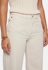 Beige weitgeschnittene Jeans aus einem weichen Baumwollmix, mit einem Knopf- und Reißverschluss, frontalen Taschen und dezenten Nähdetails.