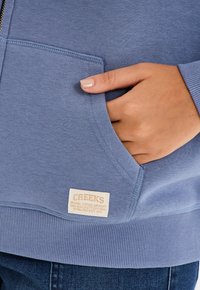 Felpa azzurra chiara con tasca frontale. Presenta un orlo e polsini a coste, e un'etichetta crema con "CREEKS" e dettagli di produzione.