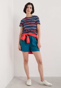 Gestreiftes T-Shirt in Marineblau, Rot und Pink; türkisfarbene Shorts; rotes Band in der Taille; weiße Sneakers; einfarbiger Hintergrund.
