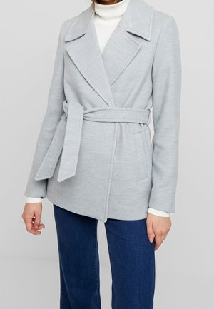 Woman wearing light gray belted coat over white turtleneck and blue jeans, standing against plain light background.