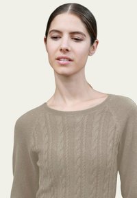 Femme aux cheveux bruns rassemblés, portant un pull en tricot torsadé beige, regardant légèrement vers le bas et sur le côté devant un fond uni.