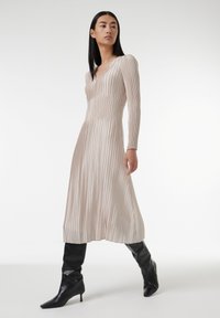 Robe plissée beige avec des manches longues, décolleté en V et design fluide. Portée avec des bottes noires arrivant au genou, mettant en avant une texture lisse.