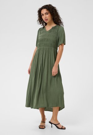 Woman with curly hair wearing a green textured midi dress with short sleeves and black heeled sandals, standing against a plain background.