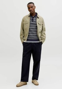 Man wearing beige jacket, patterned navy polo shirt, wide black pants, and tan suede shoes standing with hands in pockets.