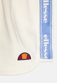 Cremefarget T-skjorte i stoff med blå sidestreker og "ellesse" tekst. Har en flerfarget brodert logo på kanten i rød, oransje og svart.
