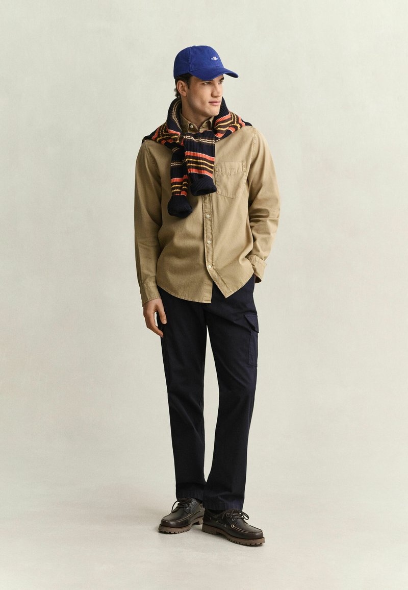 Man wearing blue cap, tan buttoned shirt with striped scarf draped over shoulders, dark pants, and brown lace-up shoes standing against plain background.