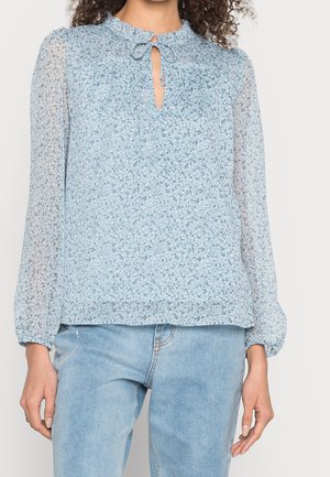 Blusa azzurra chiara in tessuto trasparente con scollatura arricciata, dettaglio con fiocco e maniche lunghe. Presenta un motivo floreale e una consistenza morbida. Abbinata a jeans in denim chiaro.