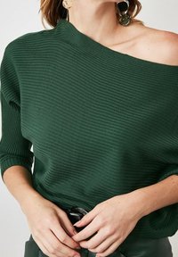 Donna che indossa un maglione a coste verde scuro, con spalle scoperte e maniche a tre quarti, grandi orecchini rotondi dal motivo marmorizzato e pantaloni scuri, con le mani unite.