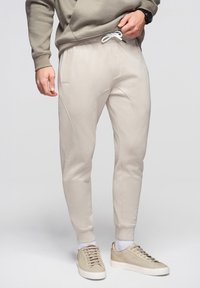 Homme ajustant un pantalon de jogging beige avec un cordon blanc, portant des baskets assorties et un sweat-shirt taupe, debout sur un fond gris clair.