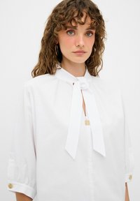Hvid bluse lavet af glat stof, med bånd om halsen, korte puffærmer og knapdetaljer ved manchetterne. Minimalistisk design.