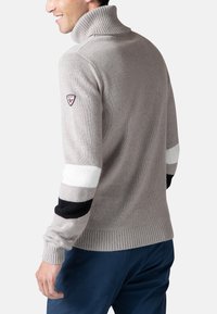 Maglione a collo alto di colore grigio chiaro, con accenti a righe nere e bianche sulle maniche. Collo e polsini a coste, logo circolare sulla manica.