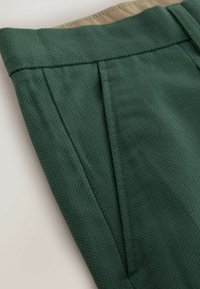 Tissu vert foncé texturé avec pli avant et doublure de ceinture beige, gros plan sur le détail d'un pantalon ajusté.