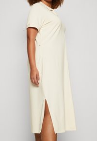 Vestido de algodón color beige claro con escote redondo, mangas cortas y aberturas laterales; corte relajado con una textura suave y detalles de diseño mínimos.