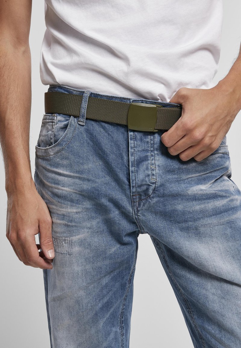 Personne portant un jean bleu délavé et une chemise blanche, ajustant une ceinture en tissu vert olive avec une boucle rectangulaire.