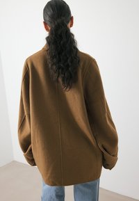 Hnedá oversized bunda zo štruktúrovanej tkaniny, s voľným strihom, spustenými ramenami a zapínaním na gombíky, nosená cez svetlomodré džínsy.