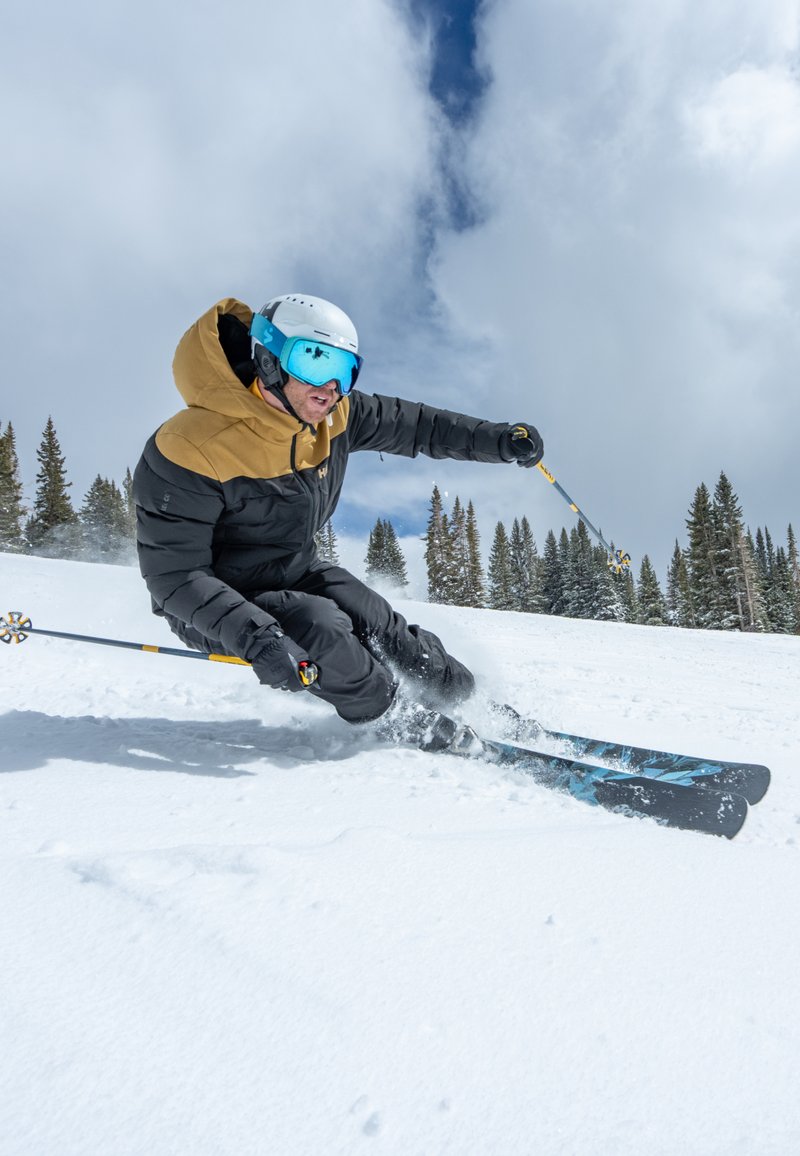 Individuele skiër op een besneeuwde helling, gekleed in een zwart en bruin jack, blauwe skibril en witte helm, gebruikmakend van zwarte ski's en gele stokken.