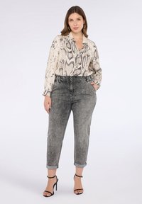 Donna che indossa una blusa a maniche lunghe con stampa di serpente, jeans grigi a vita alta arrotolati con borchie e decolletè neri con cinturino alla caviglia.