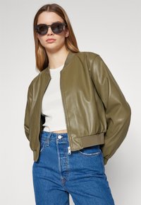Olive rohelise värvi bomber-jakk siledast kunstnahast, ribitud mansettidega, koos valge toppi ja kõrge vöökohaga siniste teksadega, ning tumedate prillidega.