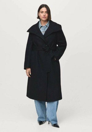 Lange zwarte wollen jas met brede kraag, ceintuur in de taille en zijzakken, gedragen over een denim shirt en wijde jeans. Glanzende zwarte schoenen.