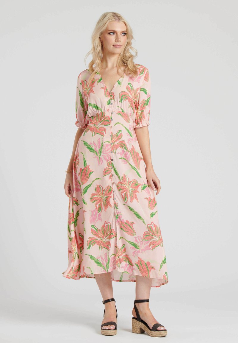 Bloemen midi-jurk in lichtroze stof met groene en roze bloemenpatronen, knoopsluiting aan de voorkant en korte pofmouwen. Een paar zwarte wedgesandalen.