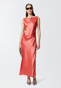 Koraljna saten haljina s dekolteom u obliku kape, prilagođenim siluetom i bočnim drapiranjem. Model drži metalik torbicu i nosi cipele s povijenim vrhom.