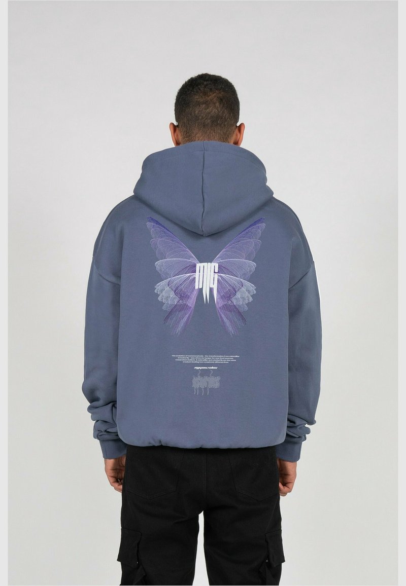 Homme portant un sweat à capuche bleu avec un papillon violet et un texte blanc imprimé au dos, debout devant un fond gris clair uni.