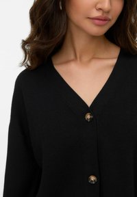 Kvinna som bär en svart V-ringad knäppbar cardigan, som visar nyckelben och läppar, med vågigt brunt hår på vit bakgrund.