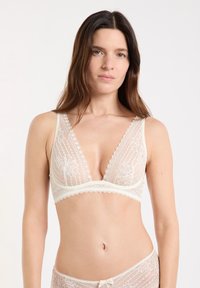 Mujer con cabello largo y castaño, vistiendo un conjunto de lencería de encaje color crema, de pie contra un fondo blanco, mirando hacia adelante con expresión neutral.