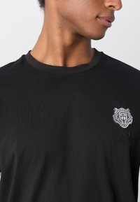 T-shirt noir en coton, avec un graphique de tigre sur le côté gauche de la poitrine et un petit logo en dessous. Design simple avec col rond.