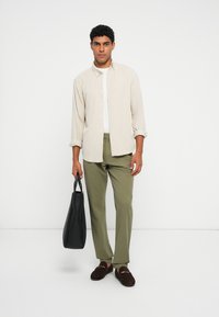 Beige skjorte med knapper over en hvid t-shirt, olivengrønne bukser, mørkebrune loafers og holder en sort tote bag. Simpelt design.