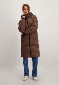 Bruine lange pufferjack met een capuchon, voorzien van een rits aan de voorkant en elastische manchetten, gedragen over blauwe jeans en witte sneakers.