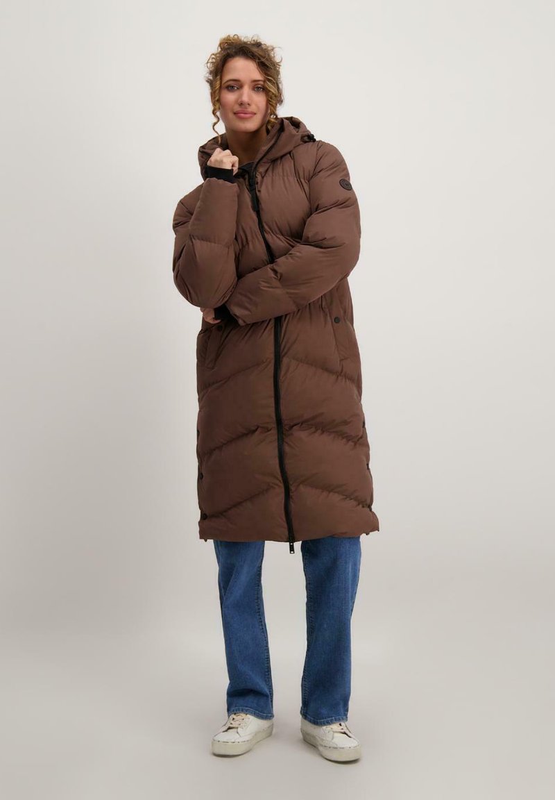 Bruine lange pufferjack met een capuchon, voorzien van een rits aan de voorkant en elastische manchetten, gedragen over blauwe jeans en witte sneakers.