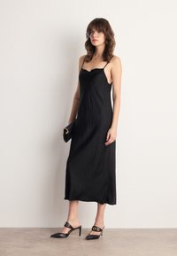 Robe de nuit en satin noir avec des bretelles fines, longueur genoux, texture lisse. Associée à des talons aiguilles noirs avec des accents ronds dorés.