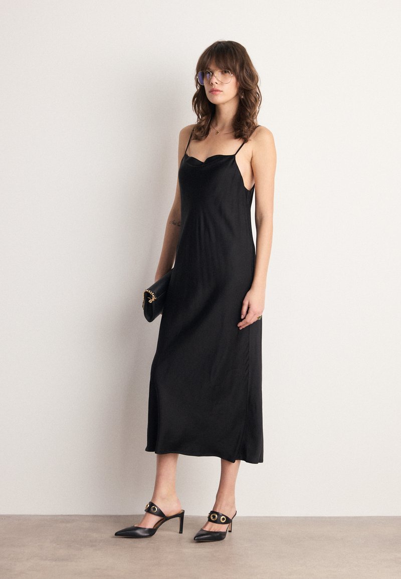 Robe de nuit en satin noir avec des bretelles fines, longueur genoux, texture lisse. Associée à des talons aiguilles noirs avec des accents ronds dorés.