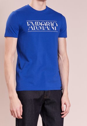 Homme portant un t-shirt Emporio Armani bleu roi avec un logo blanc, associé à un jean bleu foncé, debout devant un fond beige.