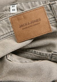 Világosszürke farmernadrág barna bőr címkével, amelyen a „JACK & JONES JEANS INTELLIGENCE” felirat szerepel, valamint egy fehér méretcímkével, amelyen a 32-es méret látható.