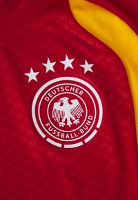 Piros sportmezes sárga részletekkel, kör alakú fehér logóval, amelyen egy sas és öt csillag látható. A szöveg: "DEUTSCHER FUSSBALL-BUND."