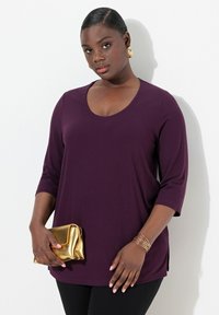 Pruimenkleurige top met een scoopneck en driekwartmouwen, gecombineerd met een gouden metallic clutch. Zwarte broek en gouden armbandjes accentueren de look.