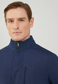 Chaqueta azul marino con cremallera y cuello alto, fabricada con tejido texturizado, con un bolsillo frontal con cremallera y detalles de costura sutiles.