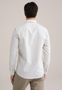 Camisa blanca de manga larga con una textura suave, puños abotonados y un dobladillo curvado, combinada con pantalones gris claro.