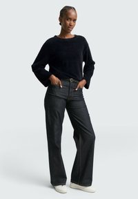 Schwarzer, fuzzie Pullover mit weiten Ärmeln, kombiniert mit dunklen, hochgeschnittenen Jeans und weißen Sneakers. Einfaches Design, neutrale Farben.