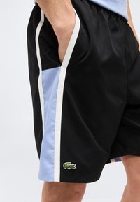 Μαύρο αθλητικό σορτς με γαλάζιες και λευκές πλαϊνές λωρίδες και πράσινο λογότυπο κροκοδείλου κοντά στο τελείωμα, φορεμένο με το χέρι στην τσέπη.