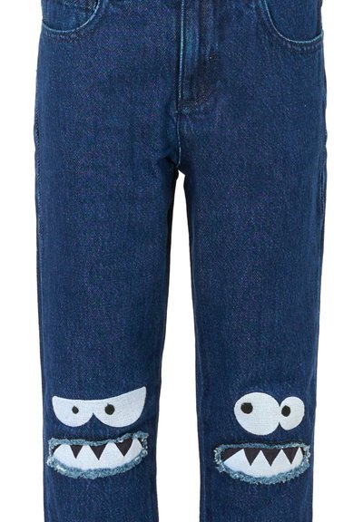 Jeans en denim bleu foncé présentant deux grands visages de monstres, au style cartoon, avec des yeux blancs et des dents déchiquetées sur les déchirures aux genoux.